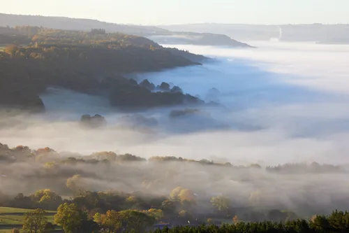 This landscape photograph captures a cloud-inversion over Curbar Edge in the Peak District, United Kingdom, at sunrise on an autumn morning. The rural nature scene in Derbyshire features layers of mist rolling through the valleys and woodland, illuminated by soft golden light from the rising sun. The atmospheric conditions typical of a cloud-inversion during autumn are prominent, with patches of woodland and fields partially obscured by the mist. Curbar Edge is a well-known landmark in the Peak District, and it enhances the natural beauty of this rural location during a dramatic sunrise.